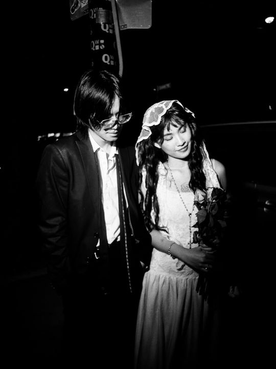 Black and white flash photograph at night: a man in a dark suit and sunglasses beside a woman in a lace veil and white dress holding roses