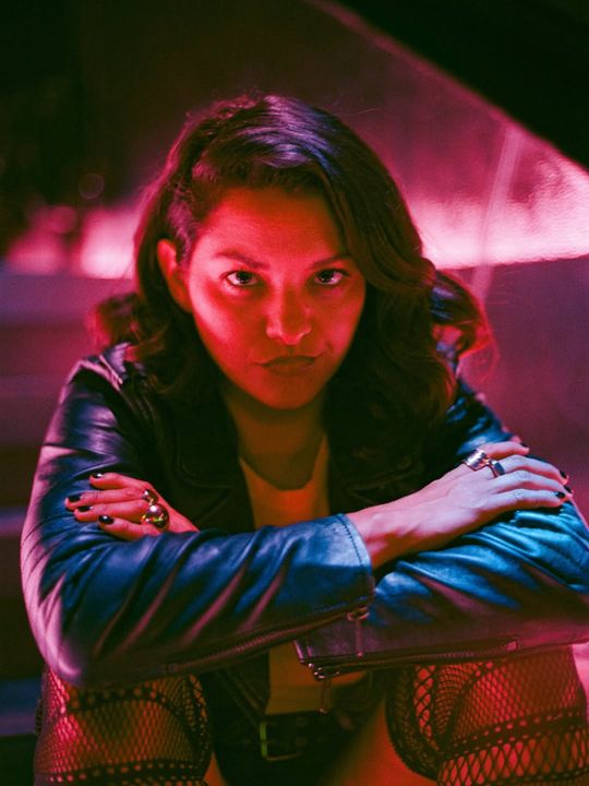 Colour portrait under red light: a woman in a leather jacket leans forward with her arms crossed, looking straight into the lens