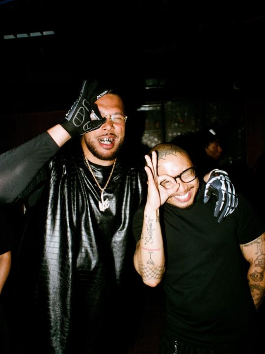 Colour flash photograph at a party: two men laughing, one in a black leather vest and gloves, the other making an OK sign over his eye